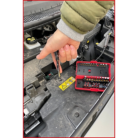 Person schraubt mit Schraubenzieher an einem Auto. Roter Werkzeugkasten mit Bits daneben. Gelbes Warnschild.